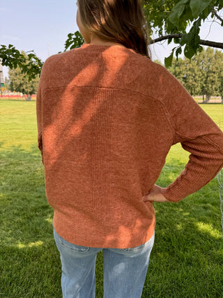 A person standing outdoors on a grassy lawn wearing a heathered rust-orange ribbed knit sweater and light blue jeans, photographed from the back.