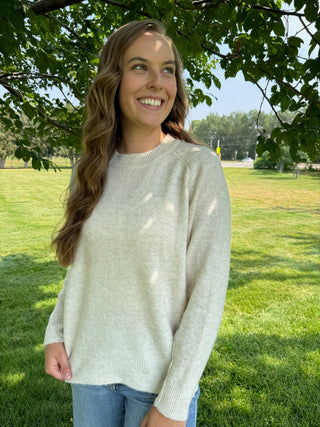 A woman smiling outdoors under a tree, wearing a light oatmeal knit crewneck sweater and blue jeans on a grassy lawn.