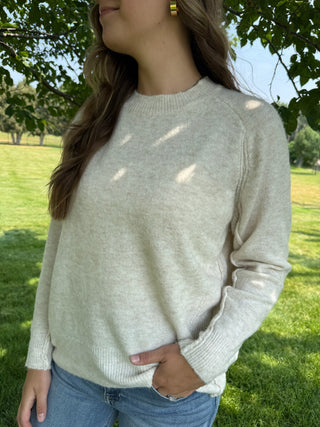 A woman wearing a light beige knit crewneck sweater and blue jeans, standing outdoors in a grassy park with trees and dappled sunlight.
