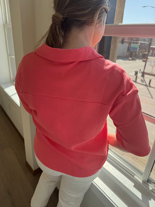 Lady standing looking out window wearing coral blouse.