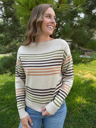 A woman smiling outdoors in front of green trees, wearing a cream knit sweater with horizontal stripes in olive green, brown, and tan, paired with blue jeans.
