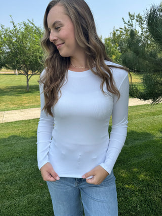A woman wearing a fitted white long-sleeve ribbed top and light blue jeans, standing outdoors on a grassy lawn with trees in the background.