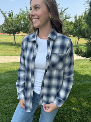 A woman wearing a blue and white plaid button-up shirt over a white t-shirt and light blue jeans, standing outdoors on a grassy lawn with trees in the background.