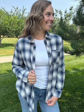 A woman wearing a blue and white plaid button-up shirt over a white t-shirt and blue jeans, standing outdoors in a grassy park area.