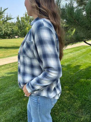 A woman wearing a blue and white plaid button-up flannel shirt and light blue jeans, standing outdoors on a grassy lawn with trees in the background.
