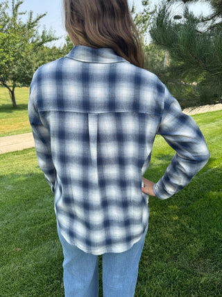 A person standing outdoors on grass, shown from the back, wearing a long-sleeve blue and white plaid button-up shirt with light blue jeans.