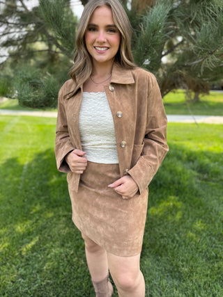 A woman standing outdoors on a grassy lawn wearing a brown suede jacket and matching skirt over a white textured top.