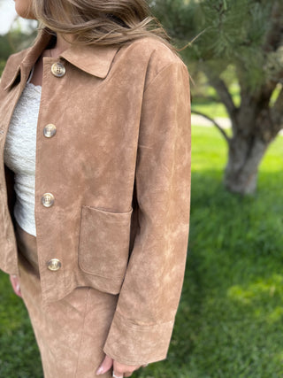 A person wearing a tan suede jacket outdoors, with a white top underneath and a matching tan skirt partially visible. The background shows a blurred green lawn and tree.