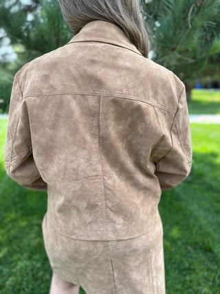 A person viewed from behind wearing a light tan suede jacket and matching skirt outdoors on a grassy lawn with trees in the background.