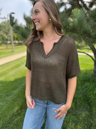 A woman wearing a short-sleeve, semi-sheer olive brown knit top with a split V-neck, standing outdoors in a park setting.