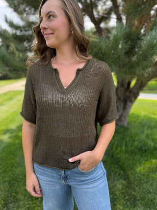 A woman wearing a short-sleeve, textured olive-brown knit top with a split V-neck, paired with light blue jeans, standing outdoors in a grassy park area.