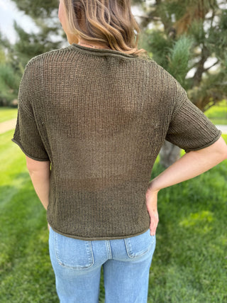 A woman viewed from behind wearing a short-sleeve textured knit top in an olive brown color, paired with light blue jeans, standing outdoors on a grassy lawn with trees in the background.