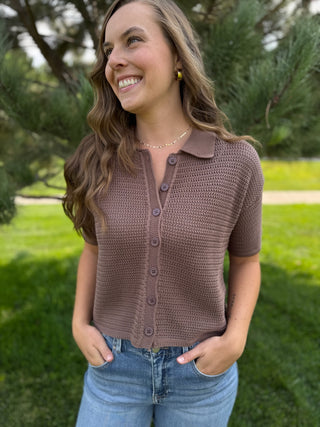 A woman wearing a brown short-sleeve knit button-up top and blue jeans, standing outdoors in a grassy park area with trees in the background.