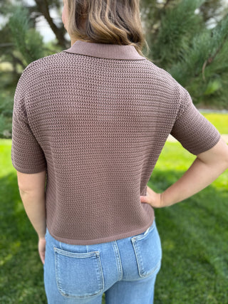 A woman viewed from behind wearing a short-sleeve textured knit polo shirt in a dusty taupe-brown color, paired with light blue jeans, standing outdoors on a grassy lawn with trees in the background.