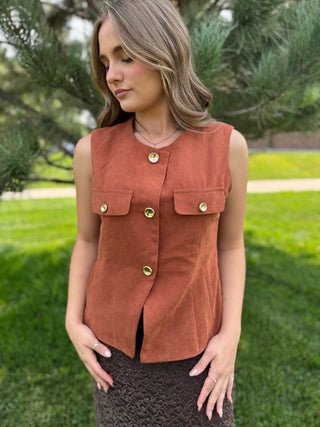 A woman wearing a sleeveless rust-brown vest with gold-tone buttons and flap pockets, standing outdoors on a grassy lawn with trees in the background.