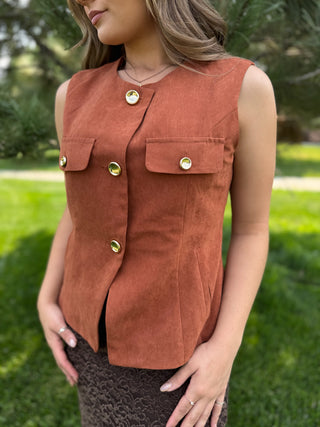 A woman wearing a sleeveless rust-brown vest with gold-tone buttons and flap chest pockets, photographed outdoors on a grassy background.