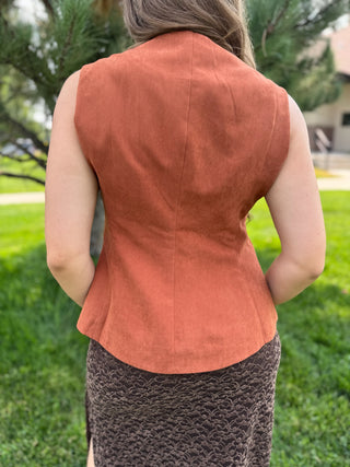 A woman viewed from behind wearing a sleeveless rust-orange vest and a textured brown skirt, standing outdoors on a grassy lawn with trees in the background.