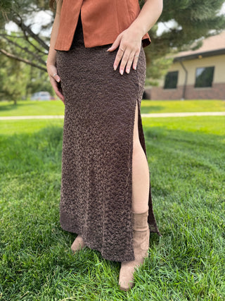 A person standing on a grassy lawn wearing a long textured brown skirt with a side slit, paired with a rust-colored sleeveless top and tan ankle boots.