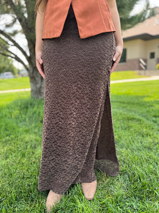 A woman standing outdoors on grass wearing a long textured brown skirt with a side slit, paired with an orange-brown sleeveless top and tan shoes.