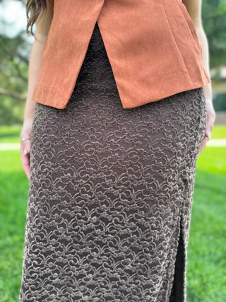 A close-up outdoor shot of a person wearing a dark brown lace skirt with a cloud-like pattern and a terracotta-colored sleeveless vest, set against a blurred green grassy background.