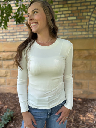 A woman wearing a fitted, long-sleeve ribbed cream-colored top and blue jeans, standing outdoors in front of a brick wall.