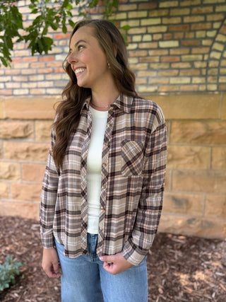A woman wearing a brown and beige plaid flannel shirt over a white top and blue jeans, standing outdoors in front of a brick wall.