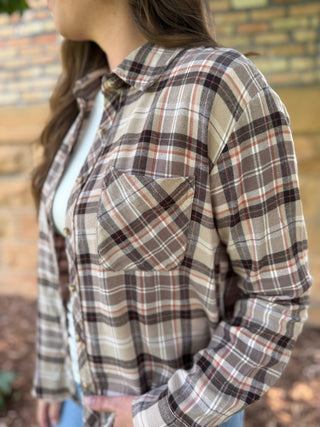 A woman wearing a long-sleeve plaid button-up shirt in beige, brown, and white tones, photographed outdoors against a blurred brick wall background.