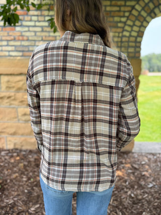 A person viewed from behind wearing a long-sleeve plaid button-up shirt in beige, brown, and white, paired with blue jeans, standing outdoors near a brick archway.