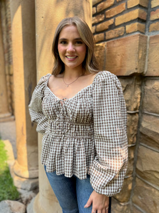 A woman wearing a brown and white gingham long-sleeve blouse with puff sleeves and a smocked bodice, paired with blue jeans, standing outdoors against a stone wall and column.