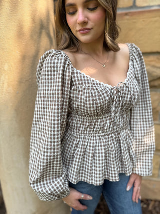 A woman wearing a brown and white gingham long-sleeve peplum top with puff sleeves and a smocked bodice, paired with blue jeans, standing against a beige stone wall.