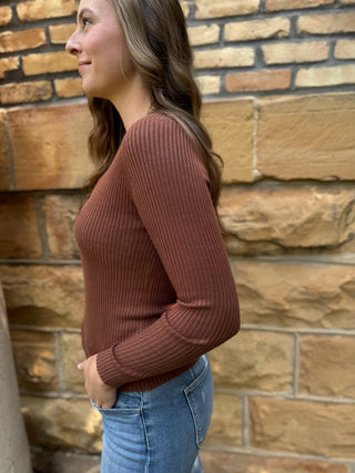 A woman in side profile wearing a rust-brown ribbed knit long-sleeve top and blue jeans, standing in front of a tan stone wall.