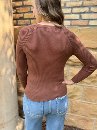 A woman wearing a ribbed long-sleeve top in a dusty terracotta brown color, shown from the back against a tan brick wall.