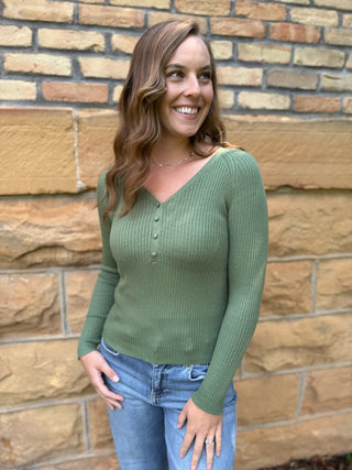 A woman smiling and wearing a ribbed sage green long-sleeve henley sweater with blue jeans, standing in front of a tan stone brick wall.