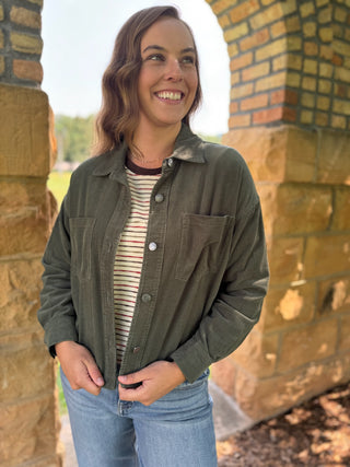 A woman smiling outdoors in front of a stone archway, wearing an olive green button-up overshirt over a striped long-sleeve top and blue jeans.
