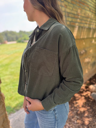 A woman wearing an olive green button-up shirt jacket with a chest pocket, paired with light blue jeans, standing outdoors next to a brick wall.