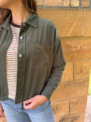 A person wearing an olive green button-up overshirt over a striped long-sleeve top and light blue jeans, standing against a tan stone wall.