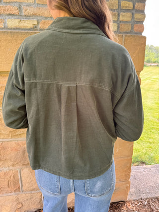 A person viewed from the back wearing an olive green cropped utility jacket with a high collar and front button closure, paired with light blue jeans, standing outdoors against a tan brick wall.