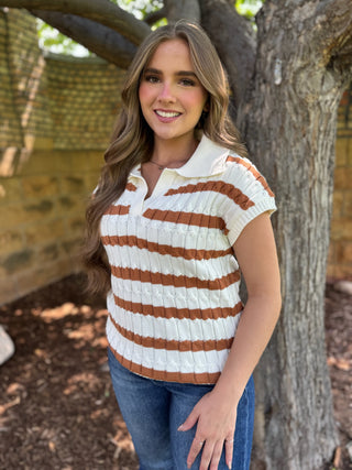 A woman wearing a short-sleeve knit polo shirt with horizontal brown and cream stripes, standing outdoors next to a tree trunk.