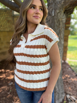 A woman wearing a short-sleeve ribbed knit polo shirt with horizontal brown and cream stripes, standing outdoors next to a tree trunk.