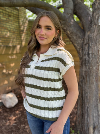 A woman wearing a short-sleeve ribbed knit polo shirt with horizontal olive green and cream stripes, standing outdoors in front of a tree trunk.