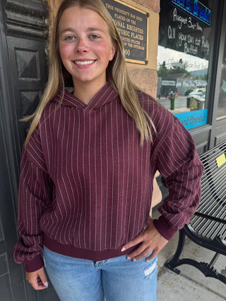 A smiling young woman with blonde hair stands outdoors in front of a storefront, wearing a burgundy hoodie with thin white vertical pinstripes and light blue jeans.