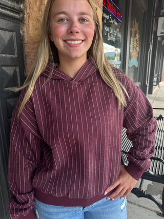 A smiling blonde woman wearing a maroon pullover hoodie with thin white vertical pinstripes and light blue jeans, standing outdoors in front of a storefront.