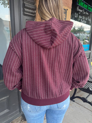 A person viewed from behind wearing a maroon pullover hoodie with thin white vertical pinstripes and a matching ribbed hem and cuffs, paired with light blue jeans in front of a storefront.