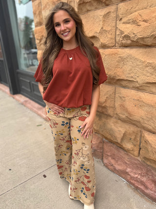 A woman stands outdoors against a tan stone wall wearing a rust-colored short-sleeve top and beige wide-leg pants with a colorful floral and butterfly print.