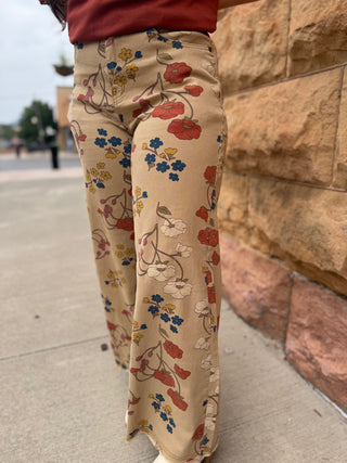 A person wearing beige wide-leg pants with a colorful floral print, standing outdoors next to a tan stone wall.