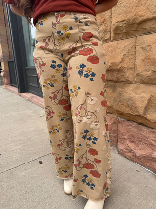 A person wearing wide-leg beige pants with a vintage floral print in red, blue, and yellow, standing on a sidewalk next to a stone wall.