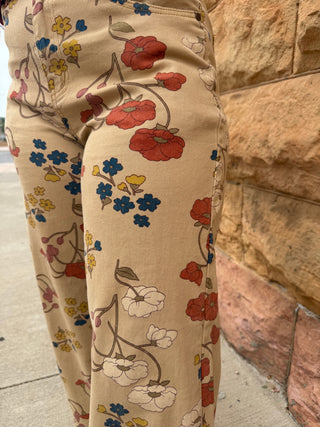 Close-up of a person wearing beige wide-leg pants with a vintage floral print in red, blue, yellow, and white, standing outdoors next to a tan stone wall.