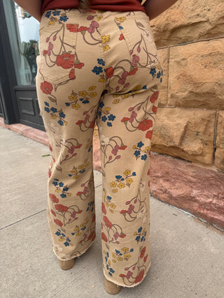 A person viewed from behind wearing beige wide-leg pants with a colorful floral vine print in red, blue, yellow, and brown, standing outdoors on a sidewalk next to a stone wall.