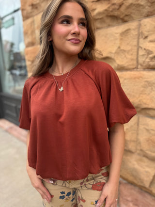 A woman wearing a rust-colored short-sleeve top and floral pants, standing outdoors against a tan stone wall.