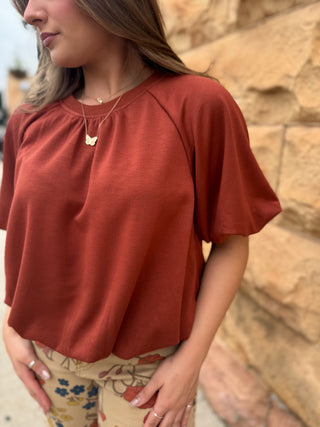 A woman wearing a rust-colored short-sleeve top with raglan sleeves and a gathered neckline, paired with beige floral pants, standing against a stone wall.
